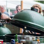 Geöffneter Big Green Egg 2XL bei einem Outdoor-Event, Gäste und Getränke im Hintergrund.