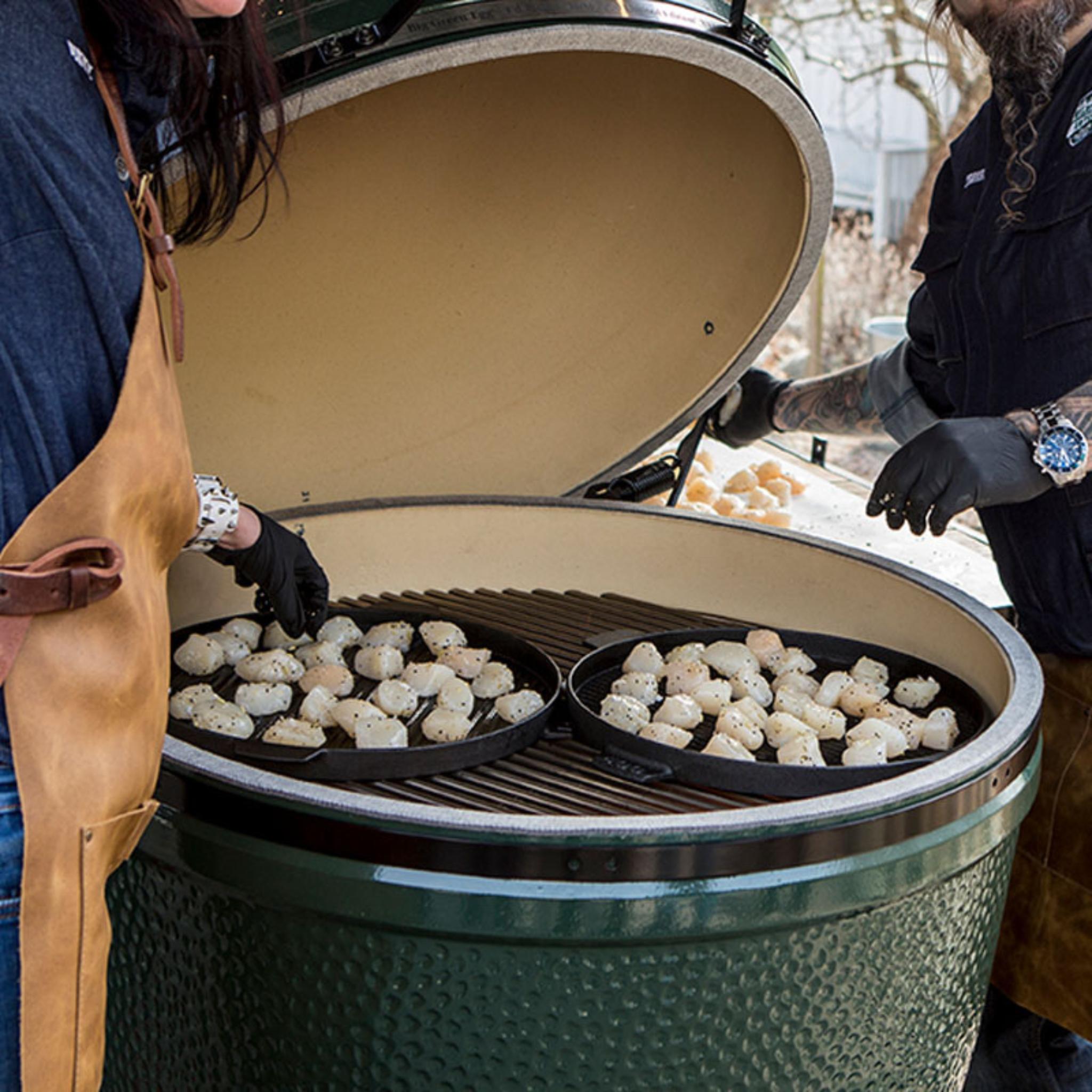 Zwei Personen grillen Jakobsmuscheln auf gusseisernen Platten im geöffneten Big Green Egg 2XL im Freien.