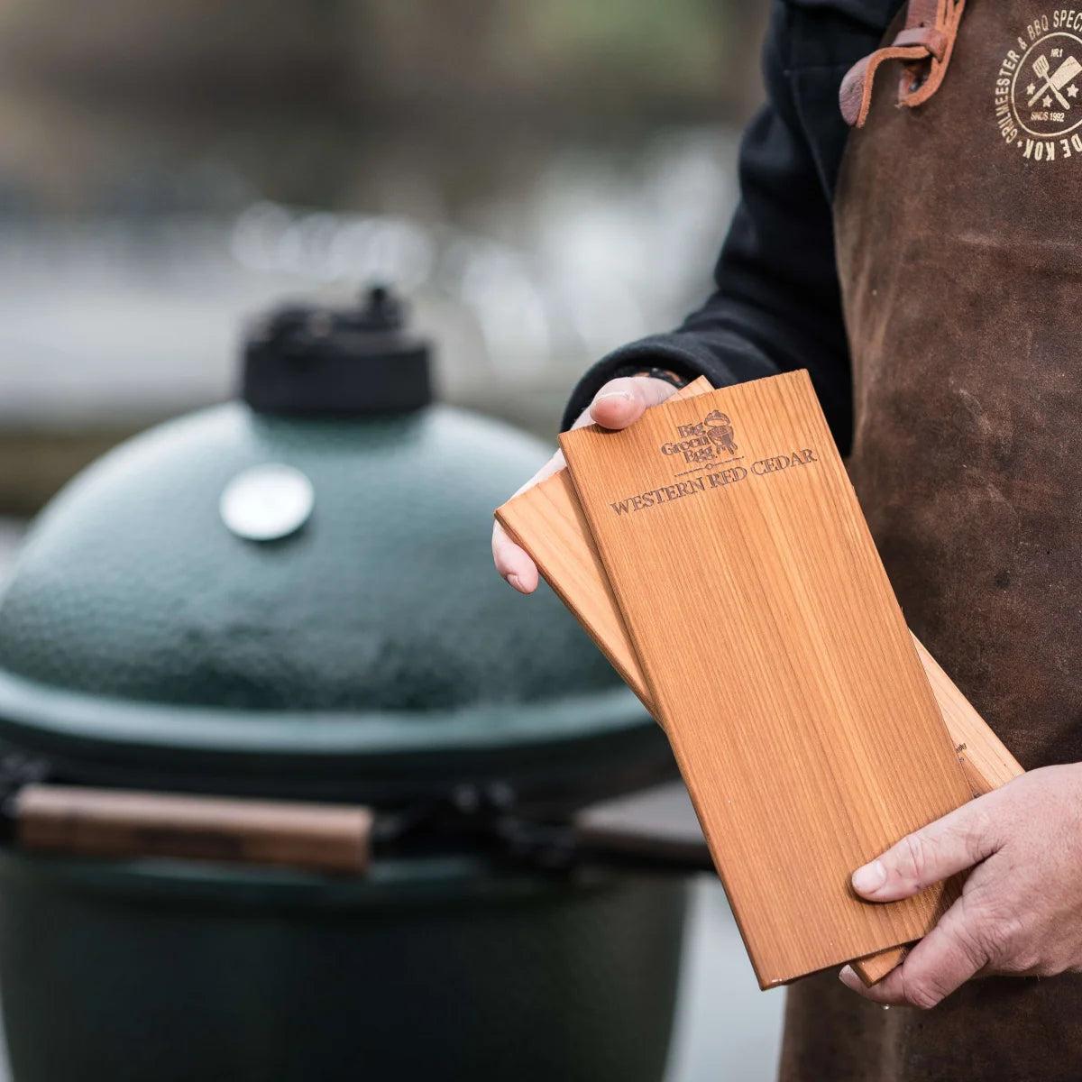 Hände halten Big Green Egg Grillplanken aus Zedernholz vor einem grünen EGG