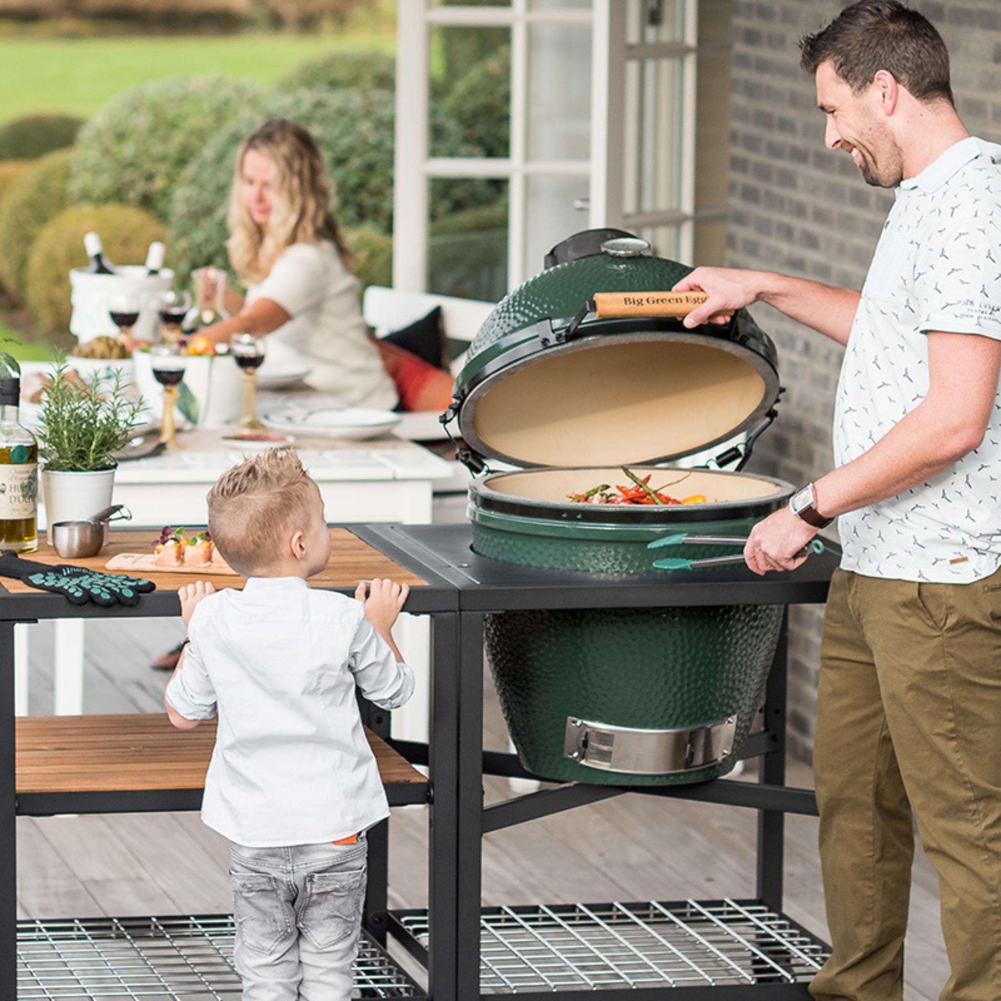 Vater öffnet den Big Green Egg Large im Tisch, Kind schaut zu, gedeckter Gartentisch im Hintergrund.