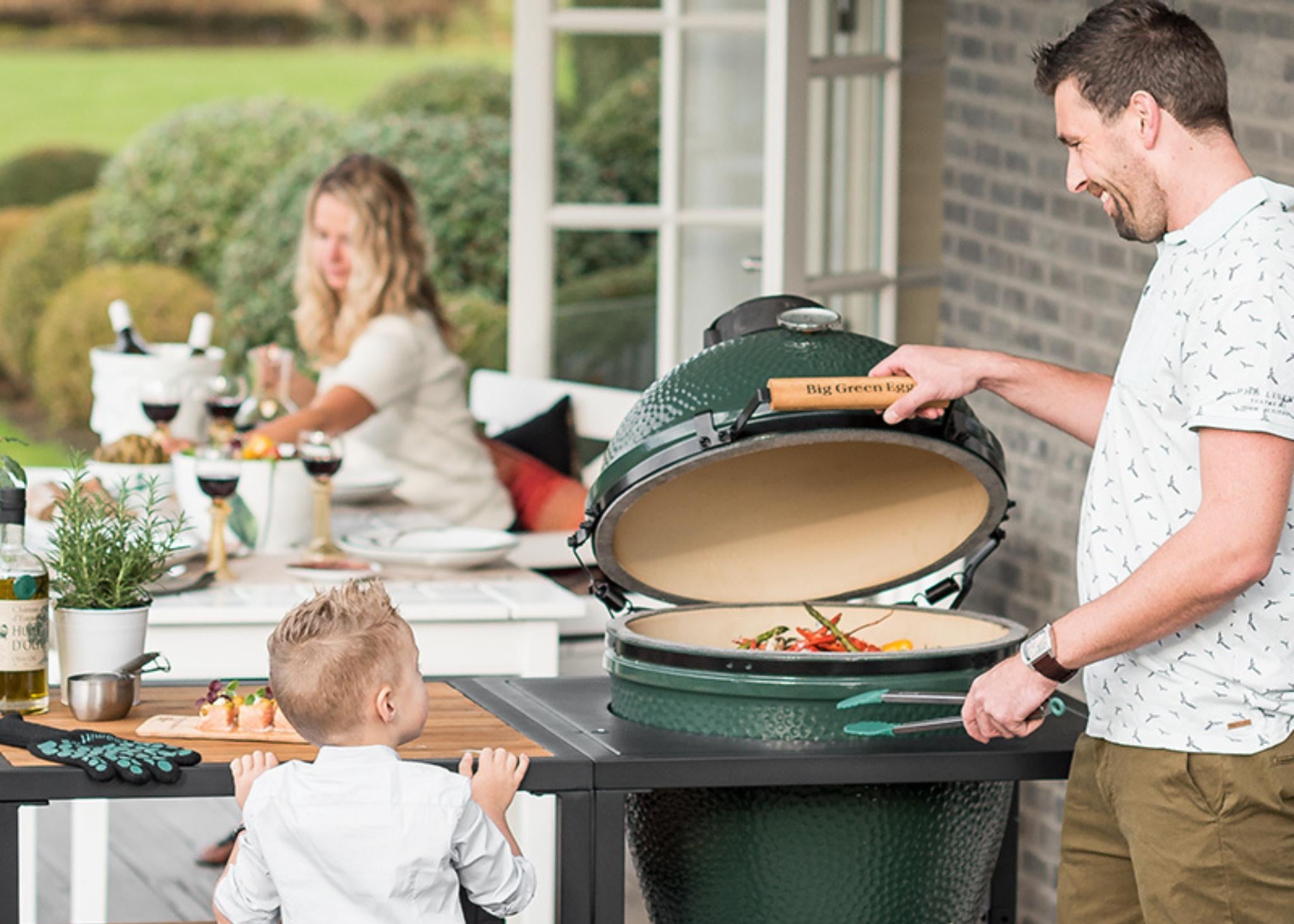 Mann am geöffneten Big Green Egg Large im Tisch mit Gemüse auf dem Rost, Kind und Gartentisch daneben.