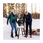 Zwei Männer stellen den Big Green Egg MiniMax auf das Portable Nest MX im verschneiten Wald