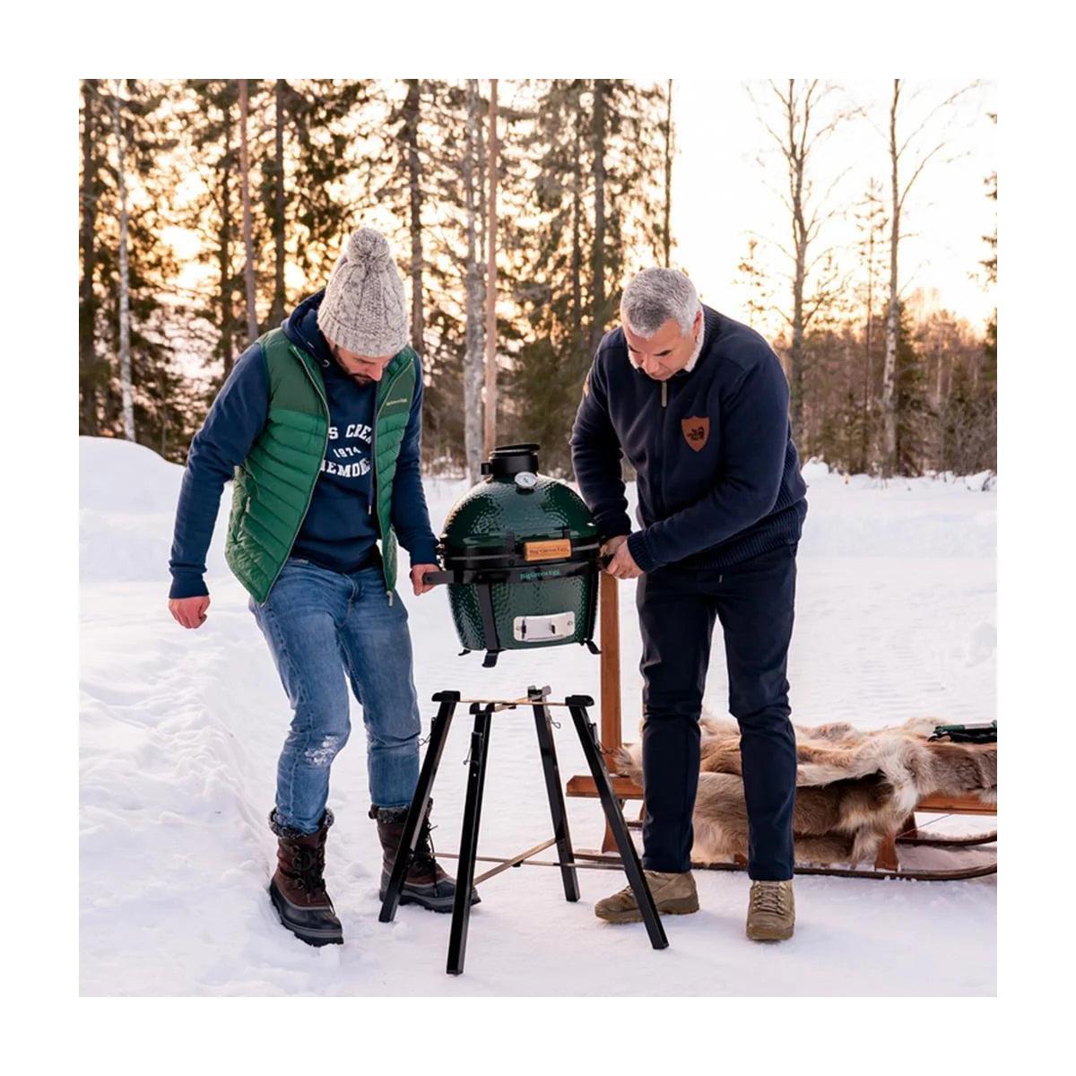 Zwei Männer stellen den Big Green Egg MiniMax auf das Portable Nest MX im verschneiten Wald