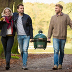 Drei Personen tragen den Big Green Egg MiniMax am Tragerahmen über einen Waldweg im Herbst
