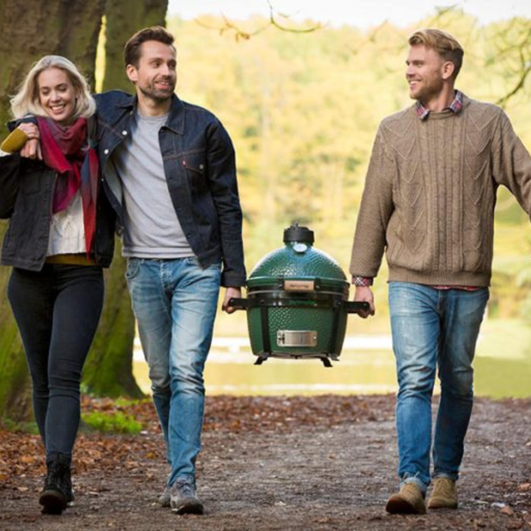 Drei Personen tragen den Big Green Egg MiniMax am Tragerahmen über einen Waldweg im Herbst