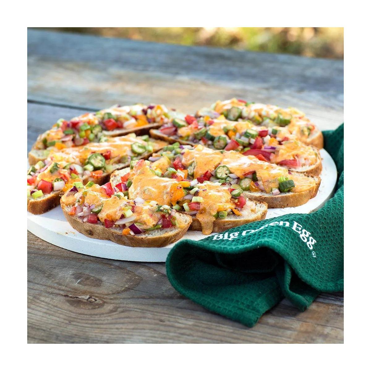 Überbackene belegte Brotscheiben mit Tomaten und Käse auf dem Big Green Egg Pizzastein, daneben grünes Tuch.