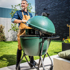 Mann mit Schürze öffnet den Deckel eines Big Green Egg XLarge auf Rollgestell im Garten