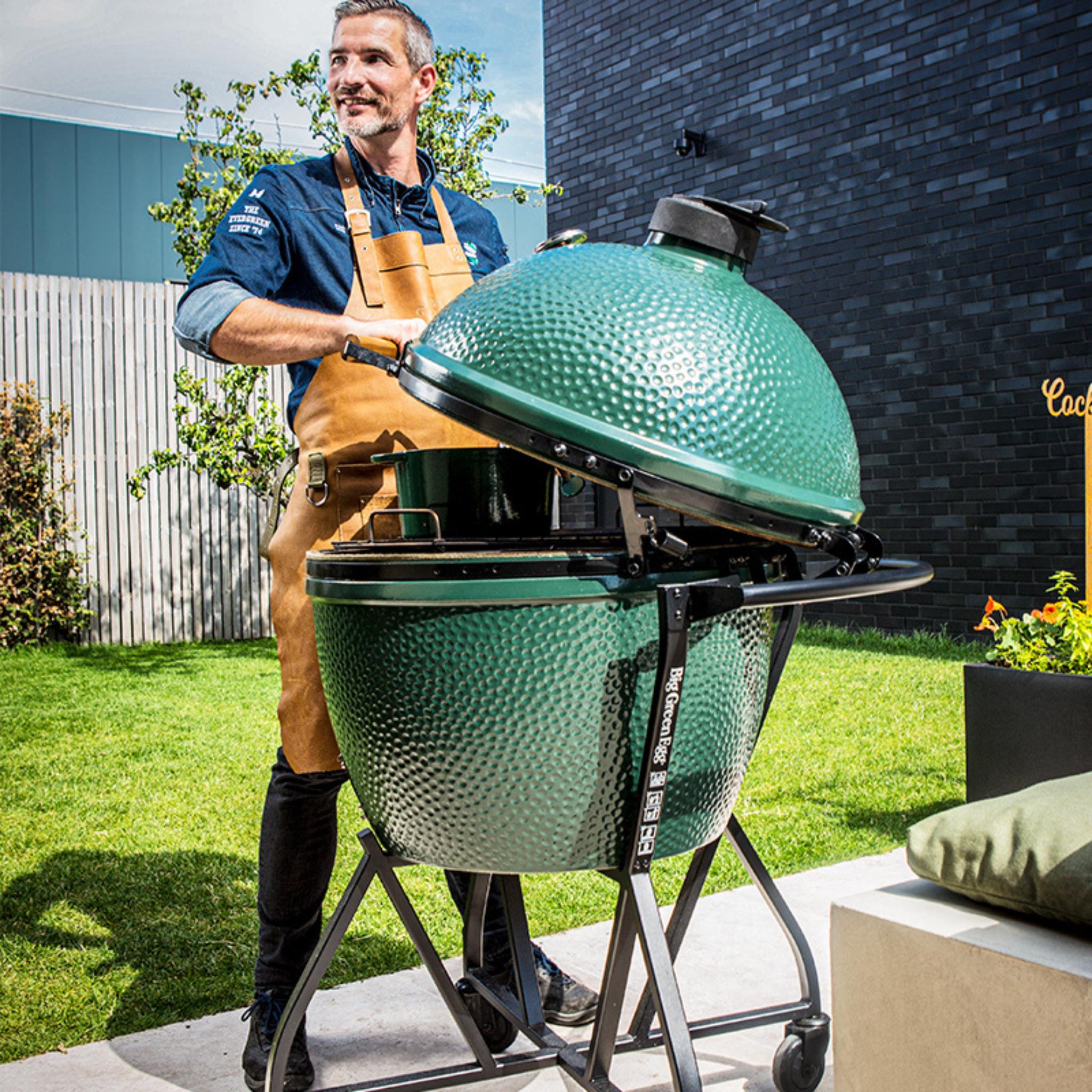 Mann mit Schürze öffnet den Deckel eines Big Green Egg XLarge auf Rollgestell im Garten
