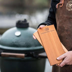 Hand hält zwei Western-Red-Cedar Grillplanken von Big Green Egg vor dem Grill