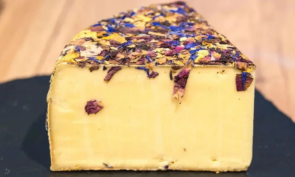 Keil eines handwerklichen Käses, die Rinde mit essbaren Blüten in lila und gelb überzogen, auf dunklem Untergrund präsentiert.