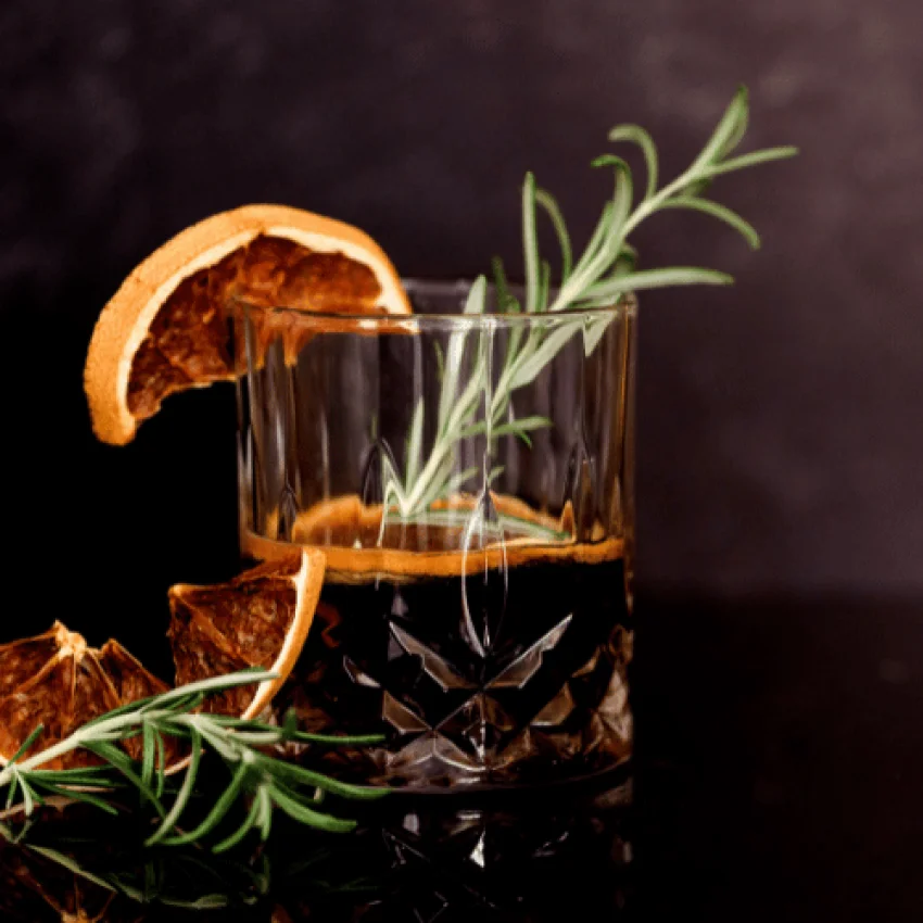 Elegantes Whisky-Glas mit bernsteinfarbener Spirituose, garniert mit frischem Rosmarin und getrockneter Orangenscheibe auf dunklem Hintergrund - miori Premium Spirituosen