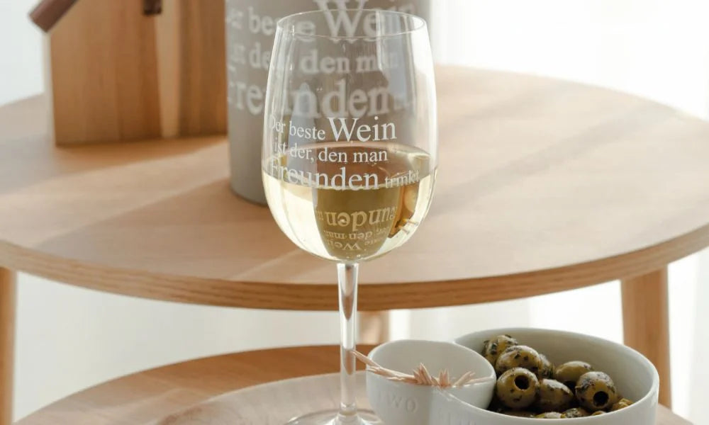 Ein Glas gekühlter Weißwein und eine Schale grüner Oliven auf einem hellen, runden Holztisch als eleganter Miori‑Aperitif.