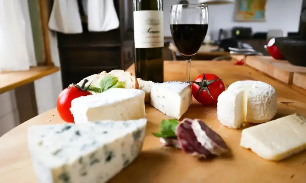 Holzbrett mit verschiedenen Käsesorten (Blauschimmel, Camembert und Hartkäse), Kirschtomaten, Brotstücken sowie Weinflasche und Weinglas im Hintergrund.