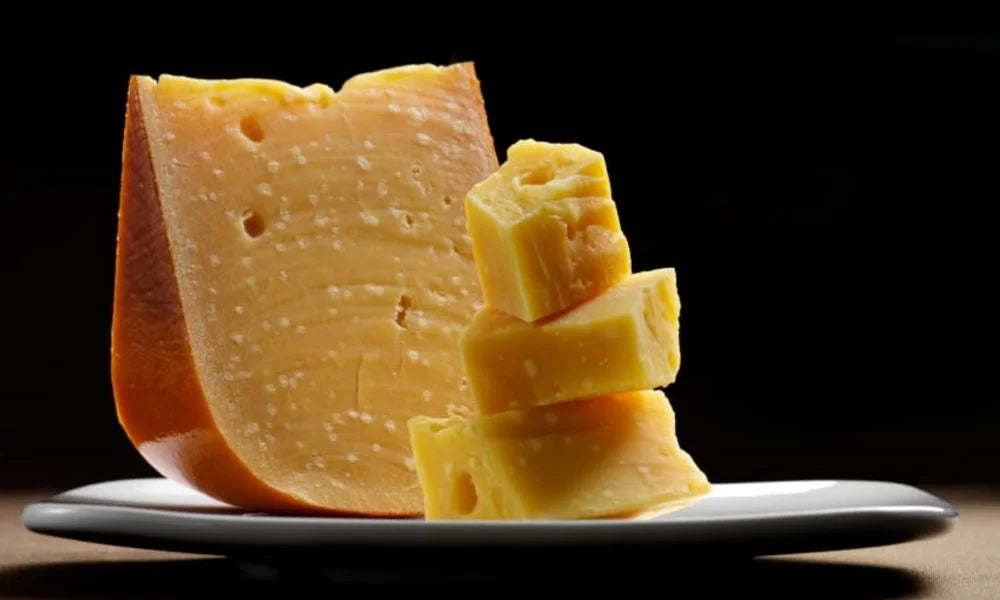 Nahaufnahme eines weißen Tellers mit Stücken und Würfeln gereiften Bauernkäse vor dunklem Hintergrund.