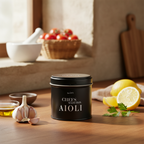 miori Aioli Dose auf Holztisch, im Hintergrund Kirschtomaten im Körbchen und Mörser, vorne Knoblauch und Zitrone