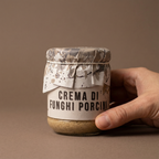 Hand hält Crema di Funghi Porcini Glas mit Papierhaube vor braunem Hintergrund