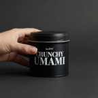 Hand greift nach Crunchy Umami Dose vor grauem Hintergrund