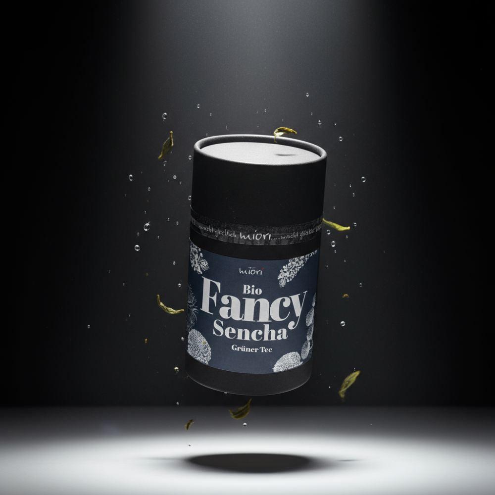 Bio Fancy Sencha Teedose mit blauem Etikett, schwebend zwischen Wassertropfen und grünen Teeblättern