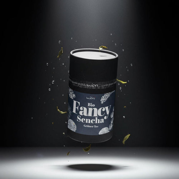 Bio Fancy Sencha Teedose mit blauem Etikett, schwebend zwischen Wassertropfen und grünen Teeblättern