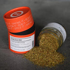 Orange Dose und gekipptes Glas Orange Ocean Rub auf grauem Stein, ausgeschüttete kräutrig-grüne Grillgewürzmischung