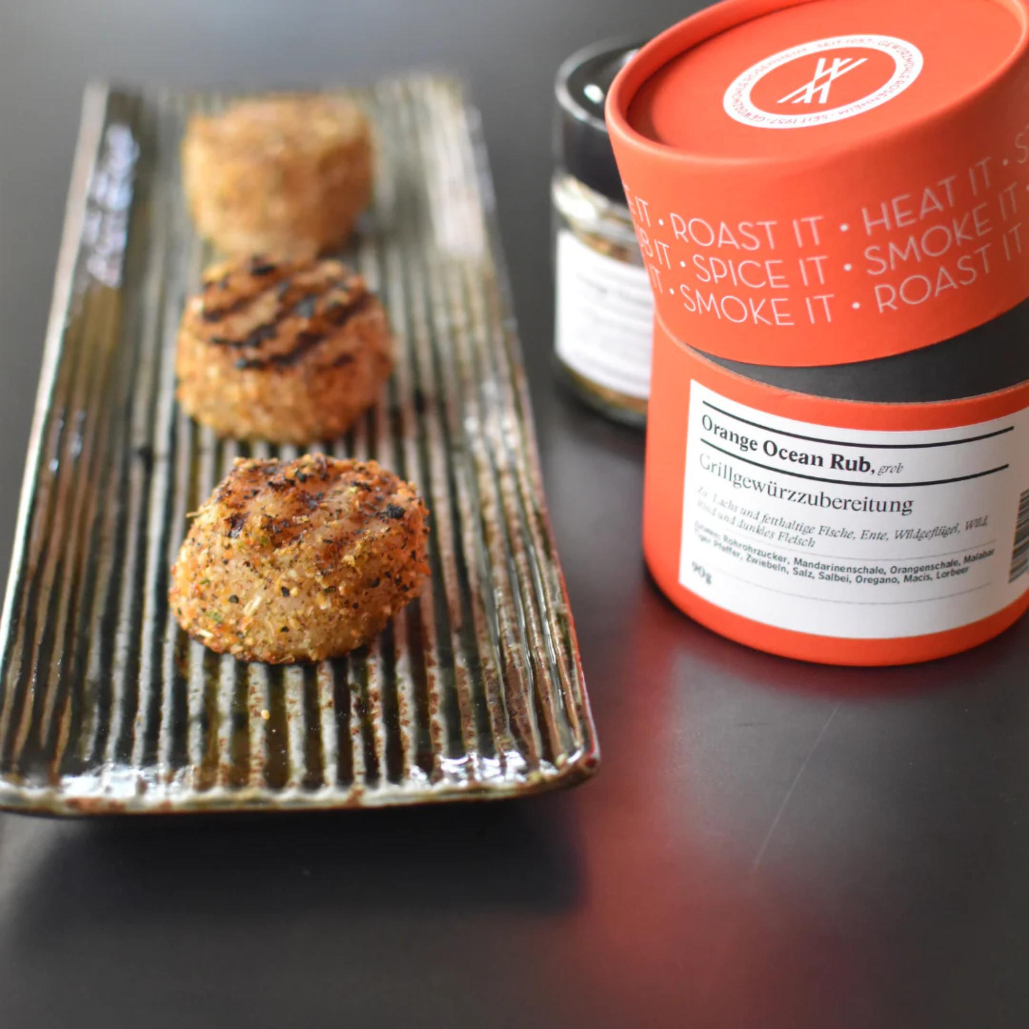 Drei gegrillte Fischfrikadellen mit Orange Ocean Rub auf gerippter Platte, orange Dose der Rosenheimer Gewürzmühle daneben