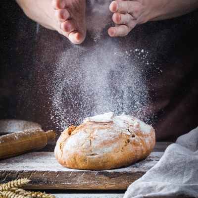 Hände bestäuben ein frisch gebackenes miori Bio  Brot mit Mehl auf einem Holzbrett.